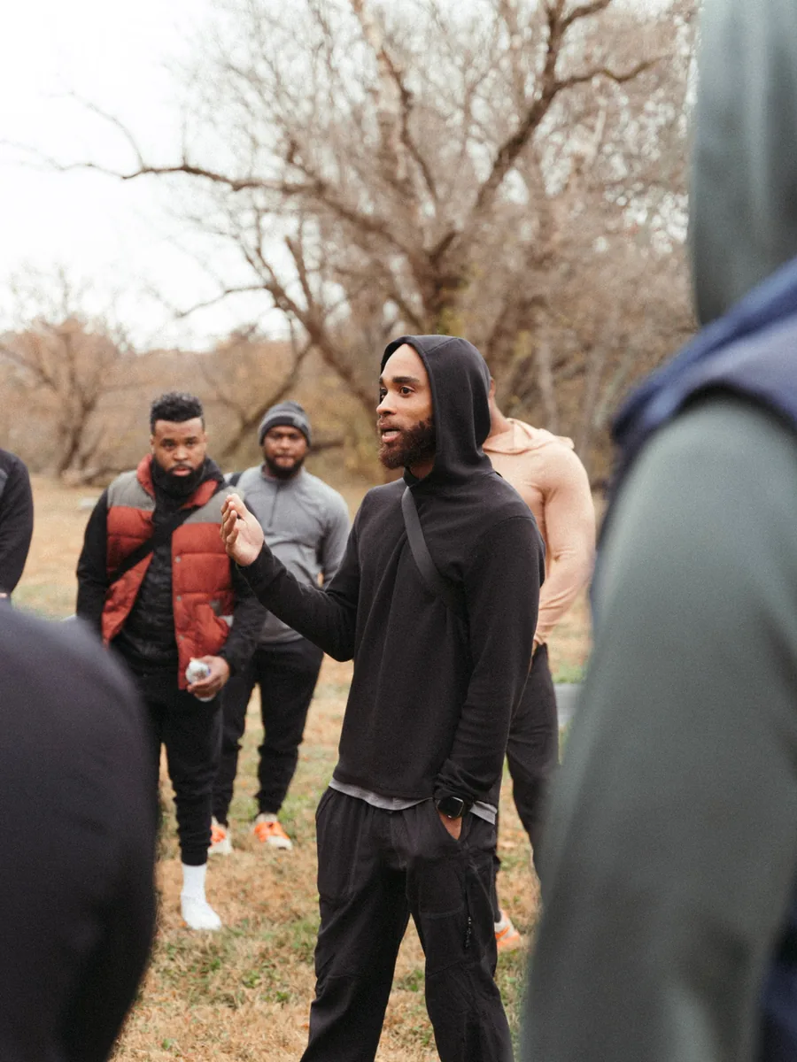 A group of men standing in a loose circle on open ground, listening while one of them speaks